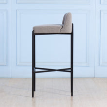 Load image into Gallery viewer, Bar stool with gray cushioned seat and black metal frame against a light blue paneled wall.