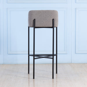 Bar stool with gray cushioned seat and black metal frame against a light blue wall.